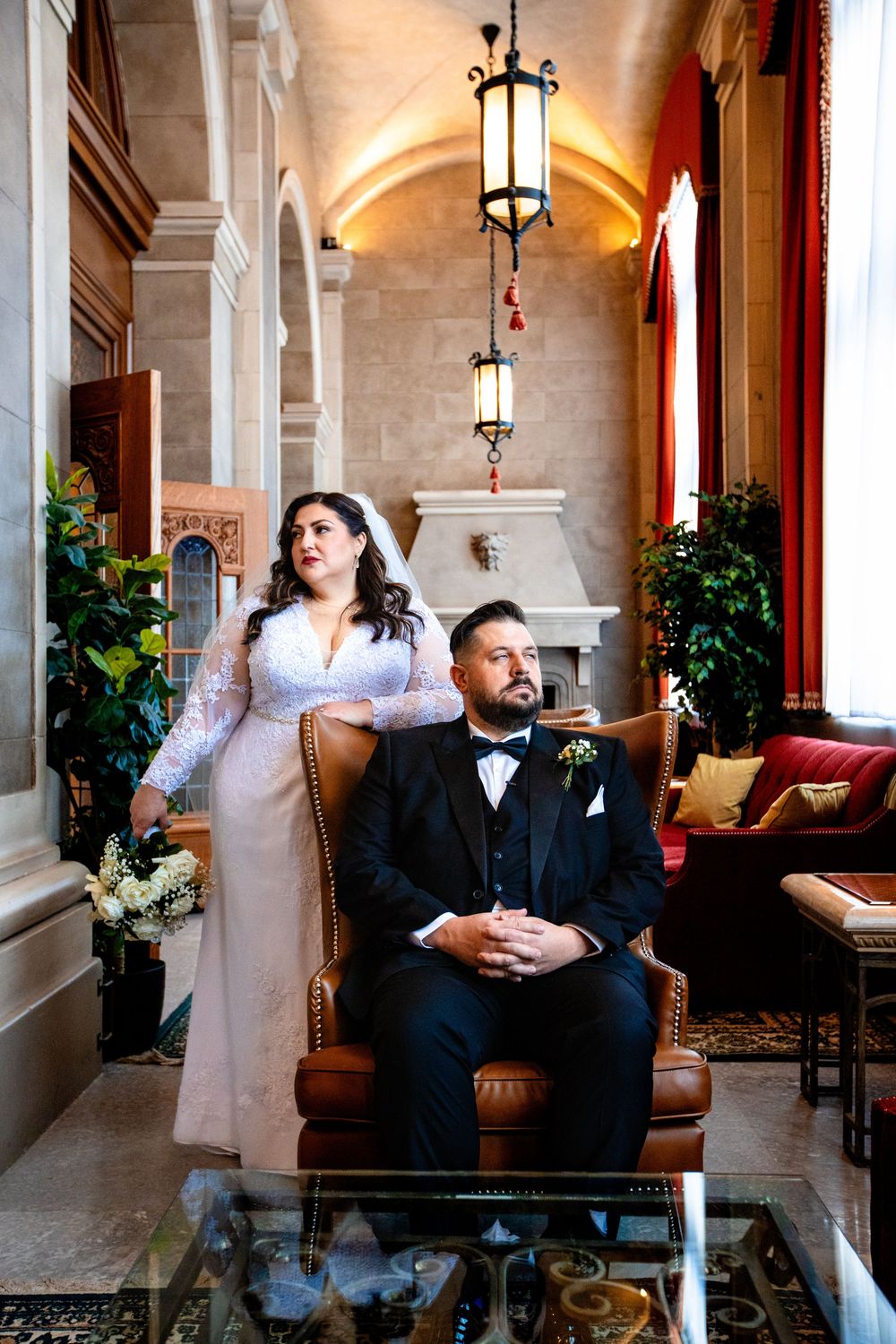 A stylish couple poses in a luxurious hotel corridor with hanging lanterns and plush furniture.
