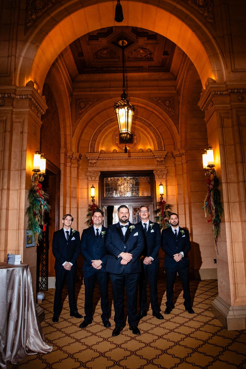 Wedding party in formal attire standing in an elegant hallway with arched ceilings and warm lighting.
