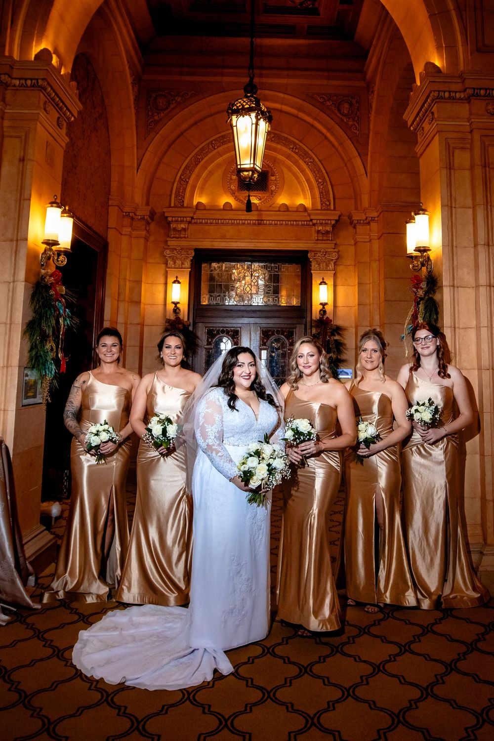 Wedding party poses in elegant venue with bridesmaids wearing champagne gold satin gowns and holding white bouquets.