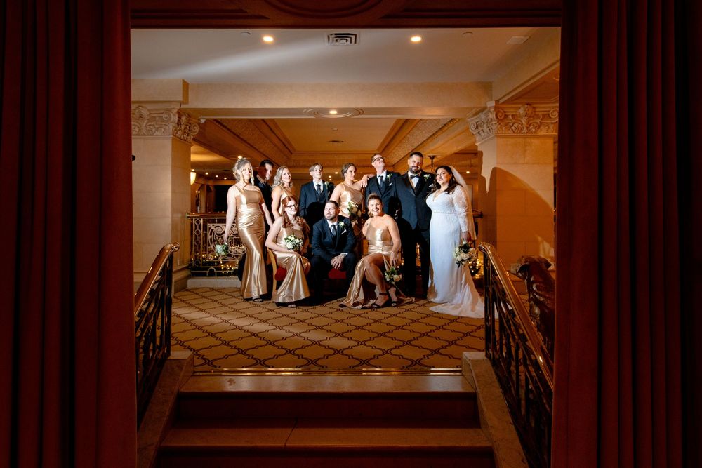 Wedding party poses on an elegant curved staircase in a luxurious hotel ballroom with warm lighting and ornate decor.