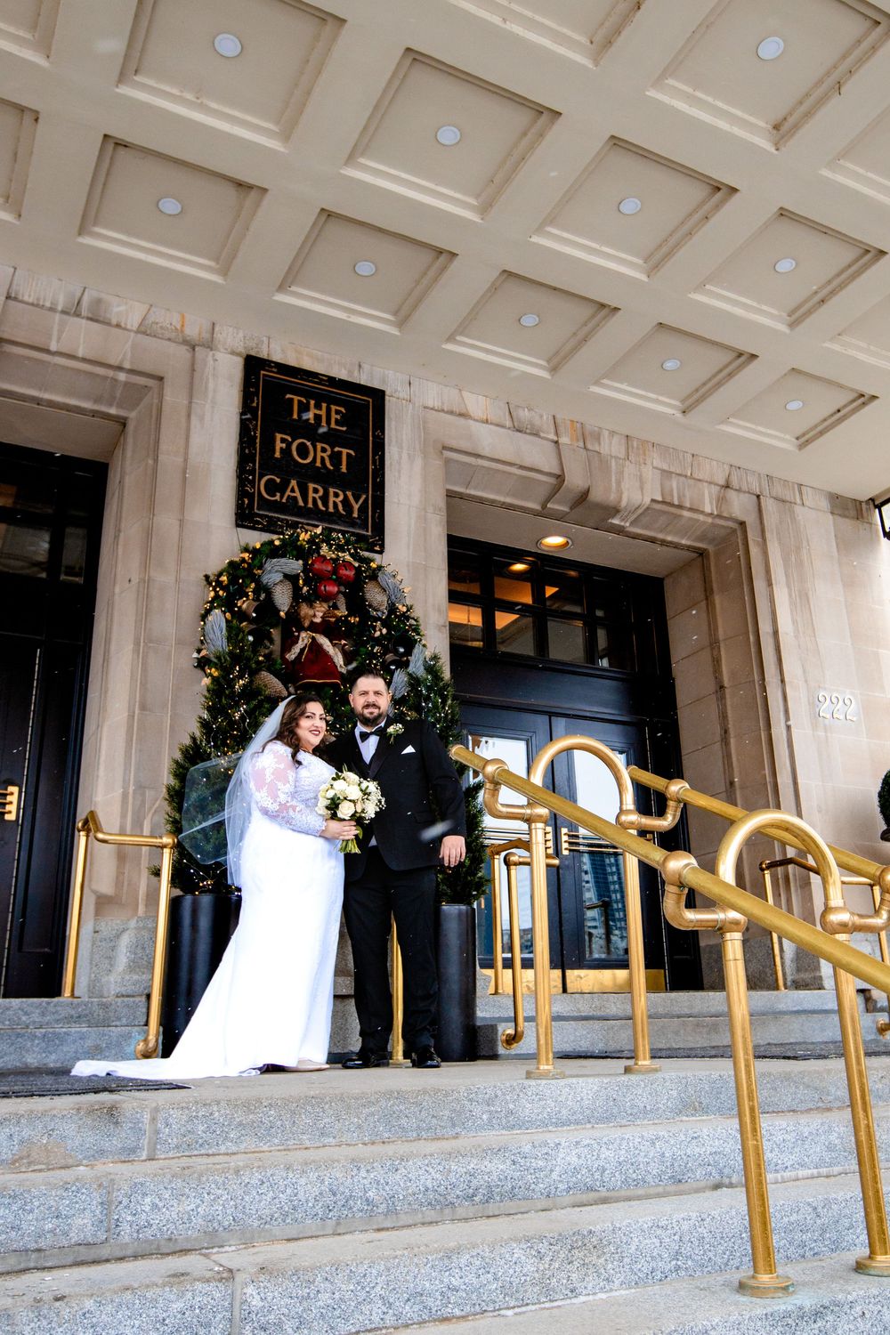 A wedding couple stands at the entrance of The Fort Garry hotel under an ornate coffered ceiling with holiday decor.