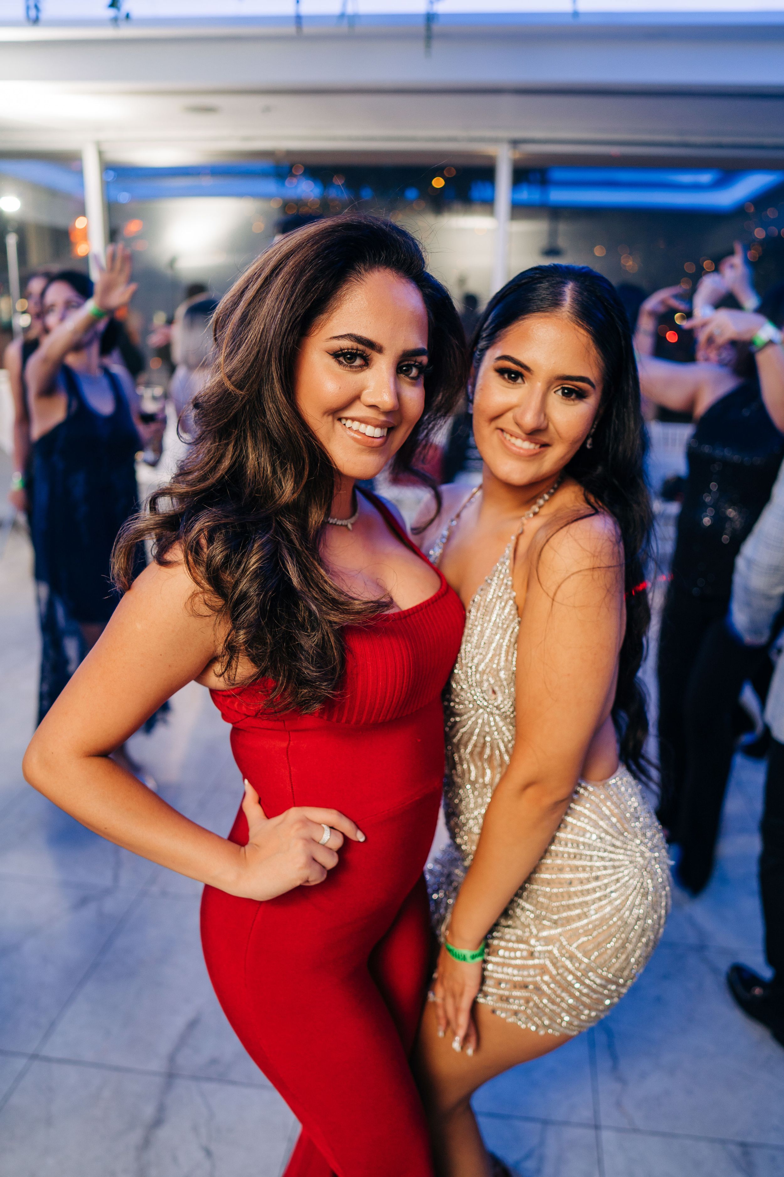 Two friends pose together at a party wearing formal dresses - one in red and one in beige with sparkles.