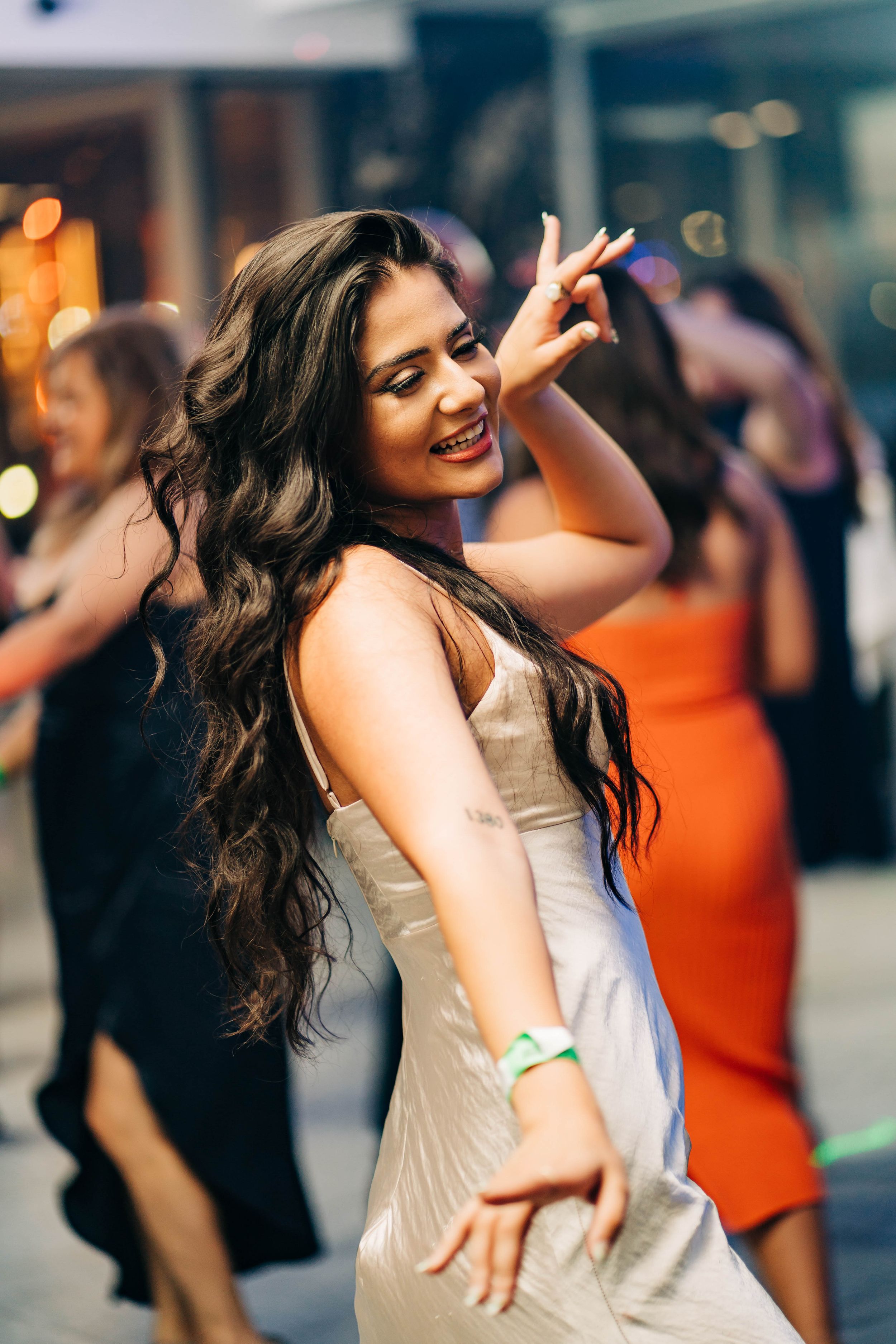 A woman with long dark hair dances joyfully in a champagne-colored dress at a party with blurred lights in background.