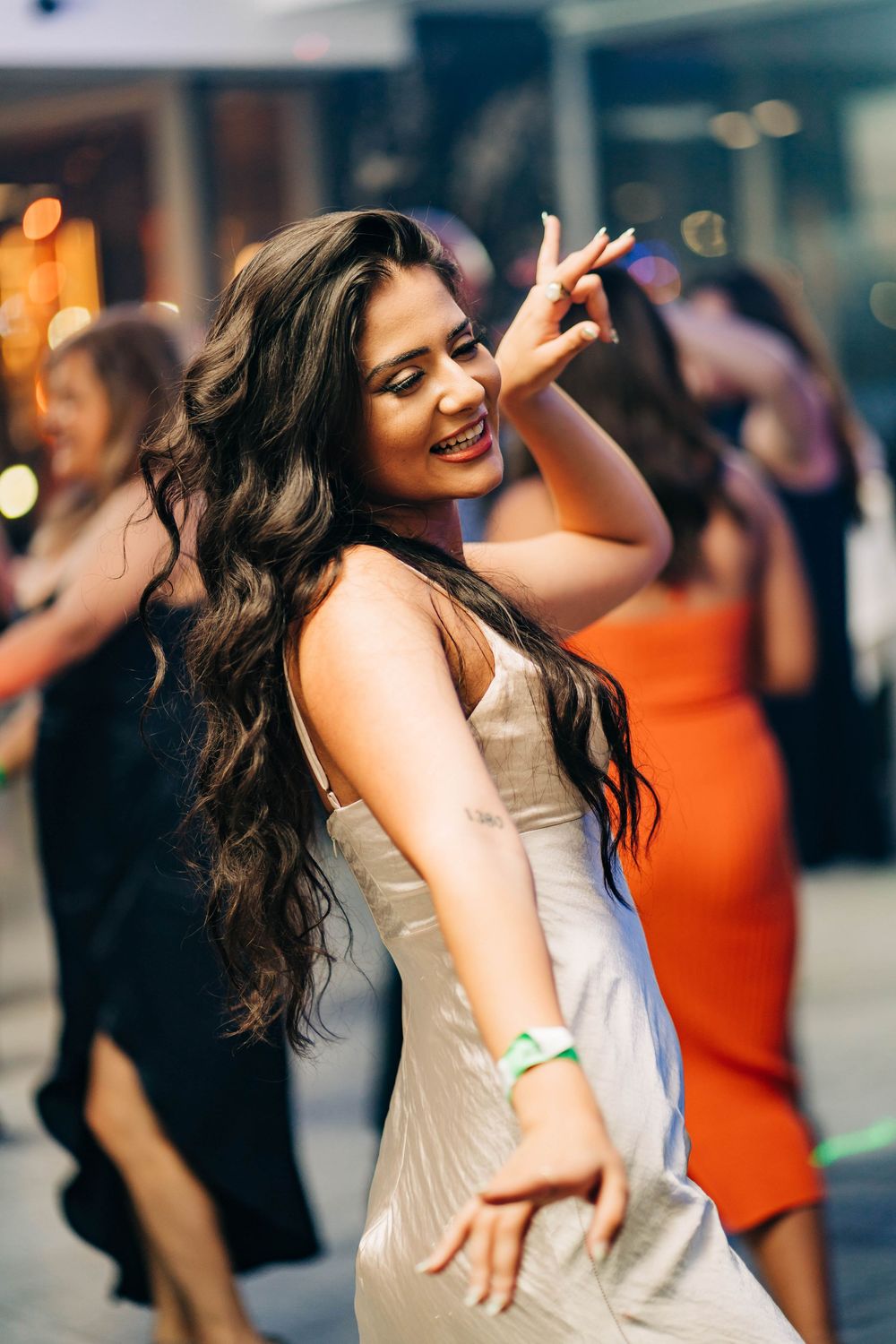 A woman with long dark hair dances joyfully in a champagne-colored dress at a party with blurred lights in background.