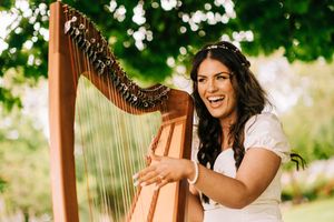 Woman in white dress performs on golden concert harp outdoors in summer garden setting.