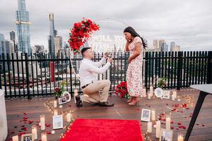 A romantic rooftop proposal setup with red balloons, candles, and a red carpet against a city skyline backdrop.