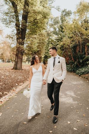 A couple in wedding attire walks along a tree-lined path during autumn with fallen leaves scattered on the ground.