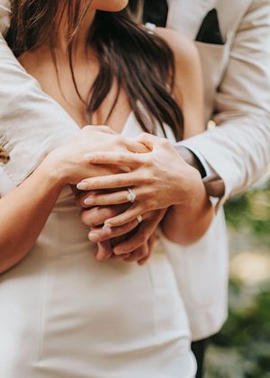 Close-up view of hands showing engagement ring and wedding band against a white dress.