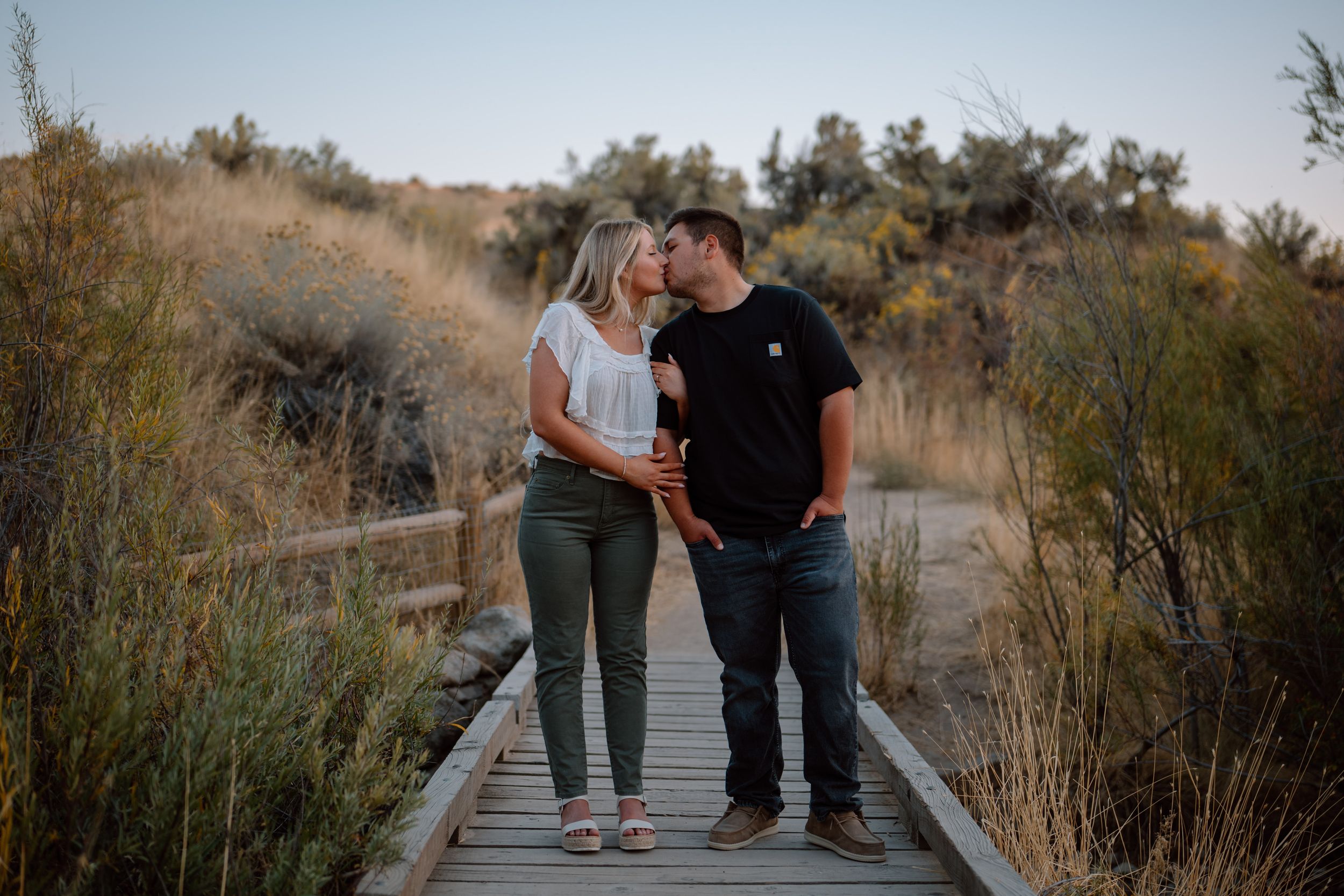 A couple shares an intimate moment while standing on a wooden boardwalk surrounded by tall grass at sunset.