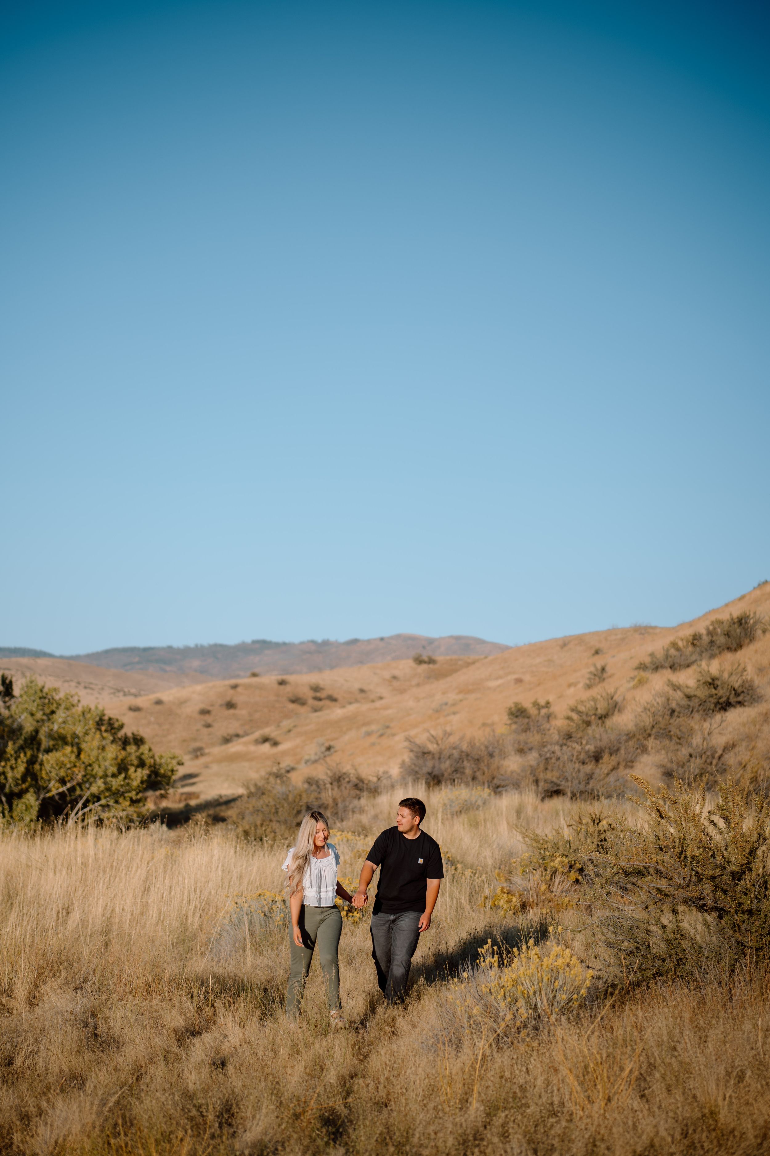 A couple walks hand in hand through golden dried grass on a hillside trail against a clear blue sky.