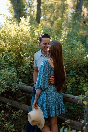 A couple shares a romantic moment in a sunlit forest path as she holds a straw hat by her side.