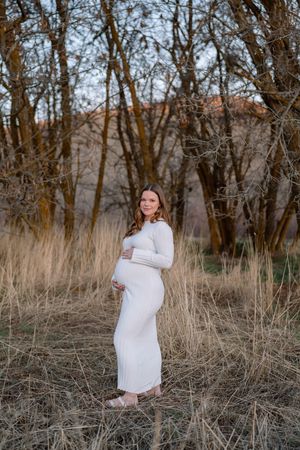 Elegant maternity photo of an expectant mother in a white dress standing among tall grass and bare winter trees.