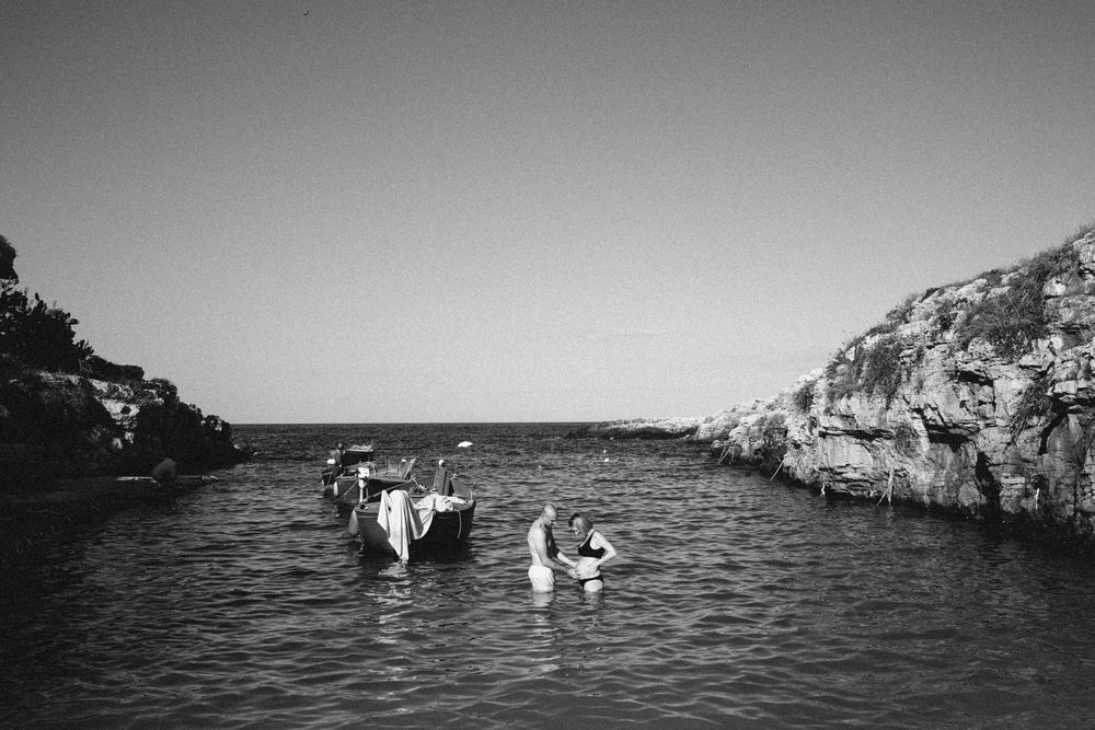 A black and white photo shows swimmers in a rocky cove surrounded by cliffs, creating a scenic Mediterranean scene.