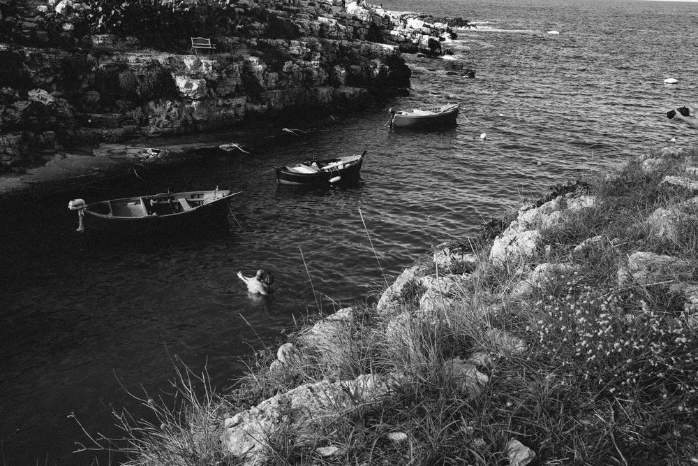 Black and white coastal scene showing small fishing boats moored along rocky Mediterranean shoreline.