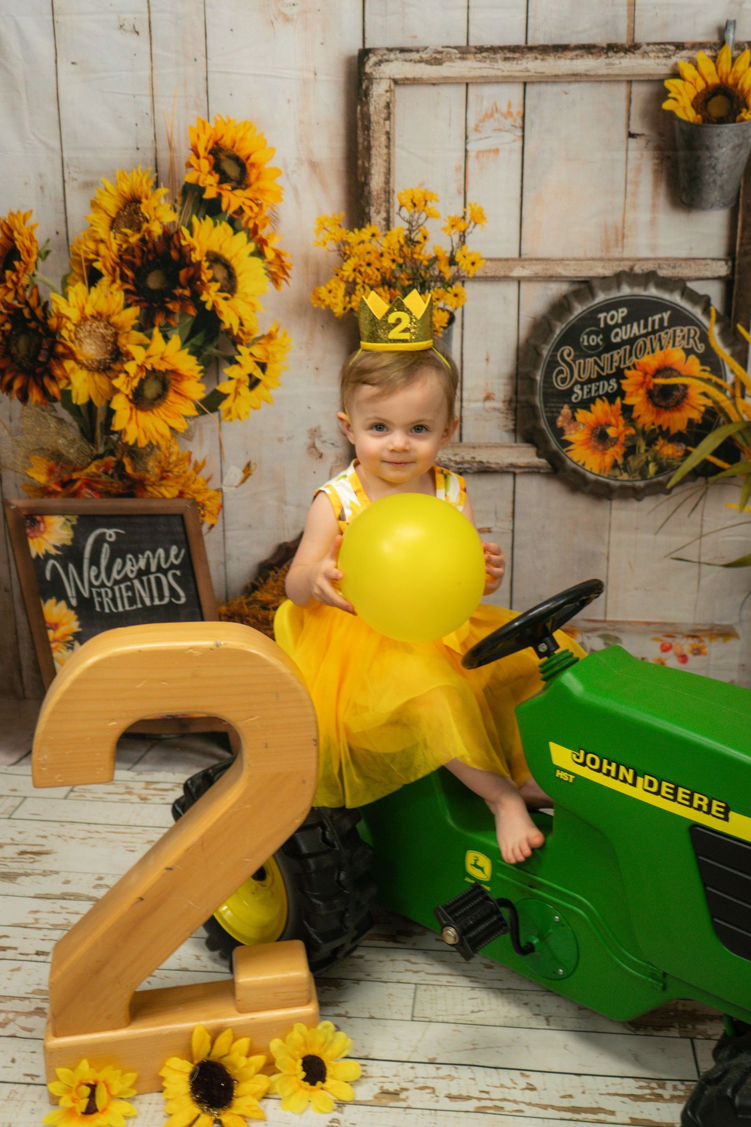 A child in yellow tulle sits on a John Deere tractor with sunflower decor and a large number 2 for a birthday photo shoot.