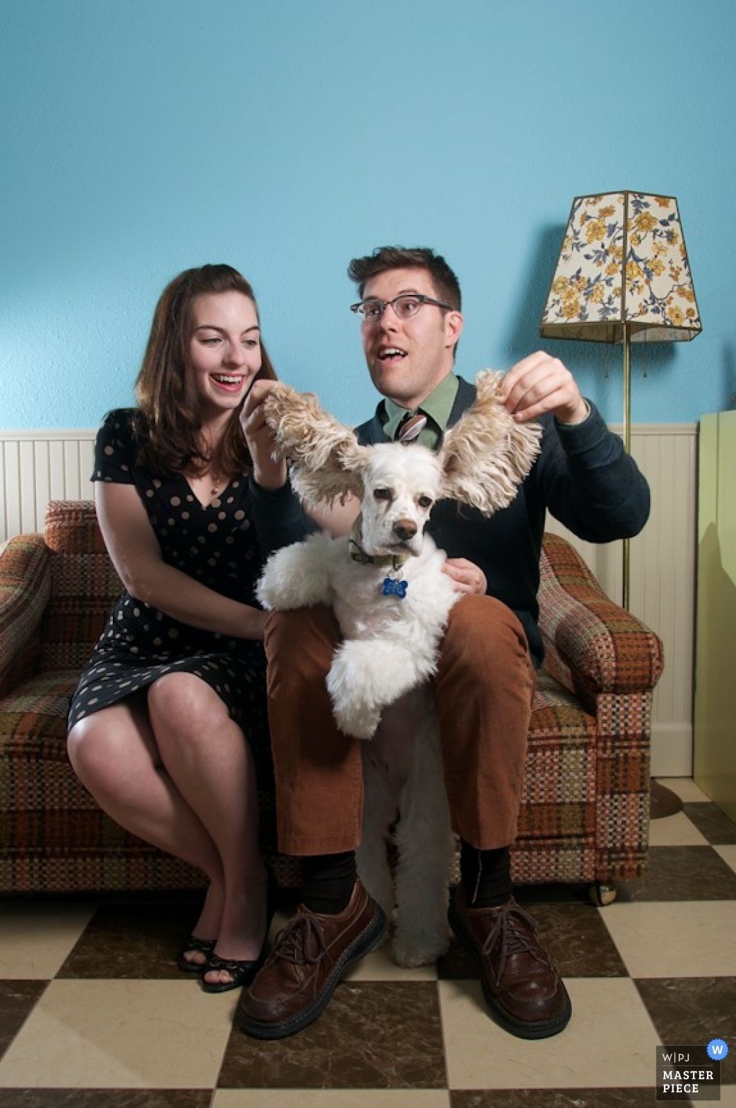 A couple sits on a vintage couch with their small white dog whose ears are being playfully lifted up in front of blue wall.