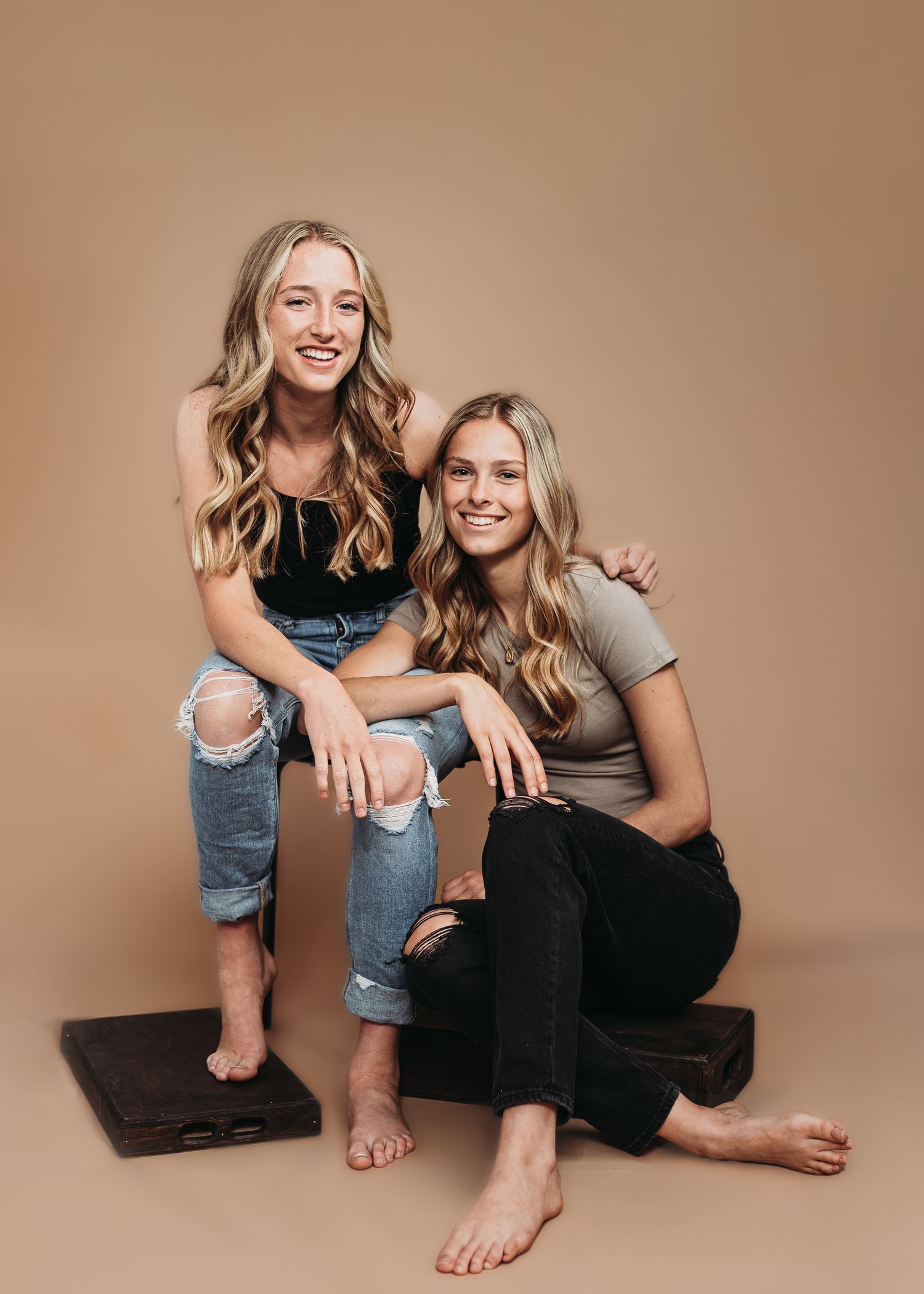 Two people in casual attire smiling and sitting together on the floor against a neutral brown backdrop.