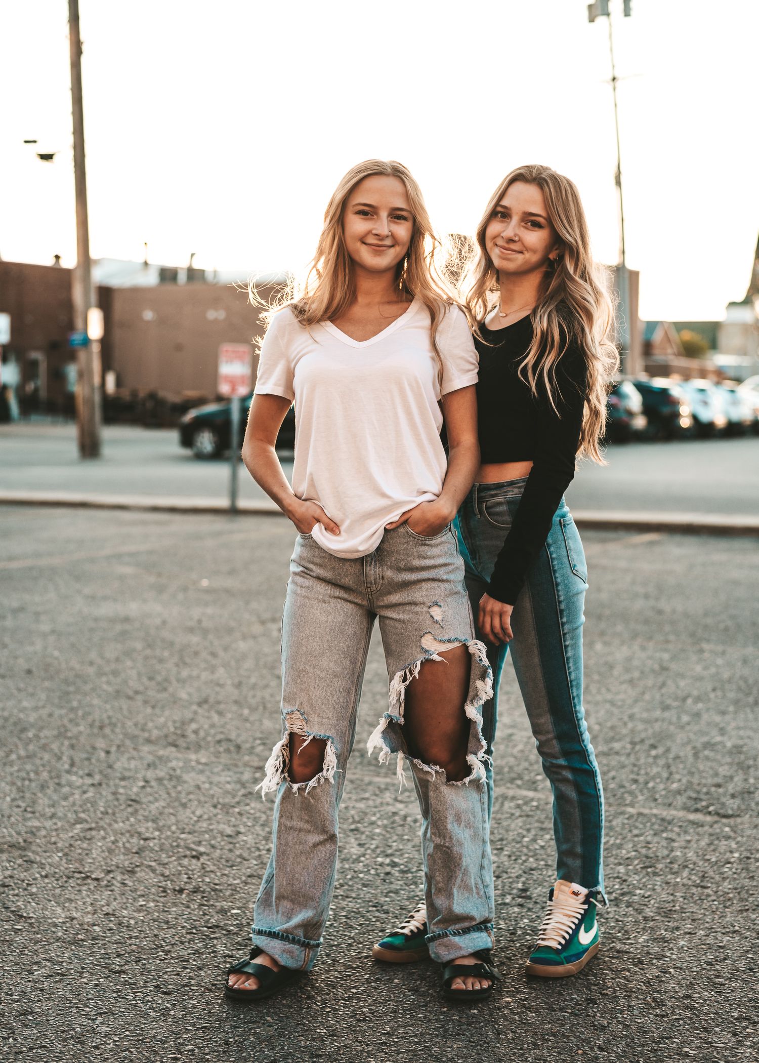 Two people in casual outfits posing together in a parking lot at sunset wearing distressed jeans and trendy tops.