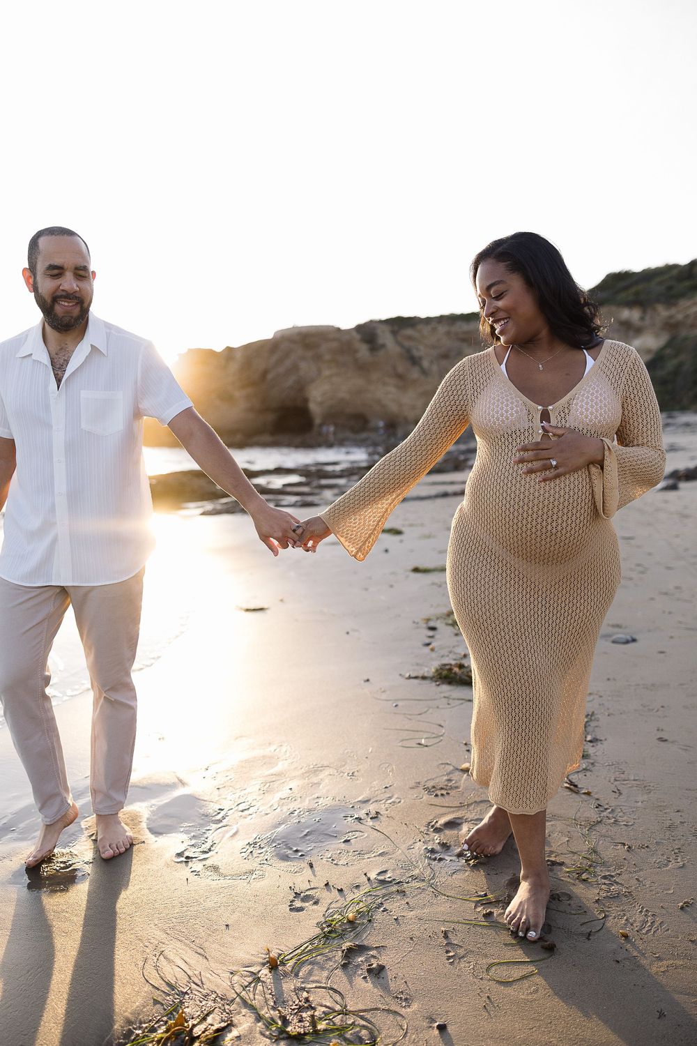 Romantic couple holds heart shape hands together at sunset on sandy beach in neutral beige and white outfits.