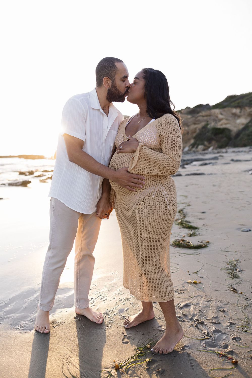 A loving couple embraces on a beach at sunset, with the expectant mother wearing a cream knit dress.