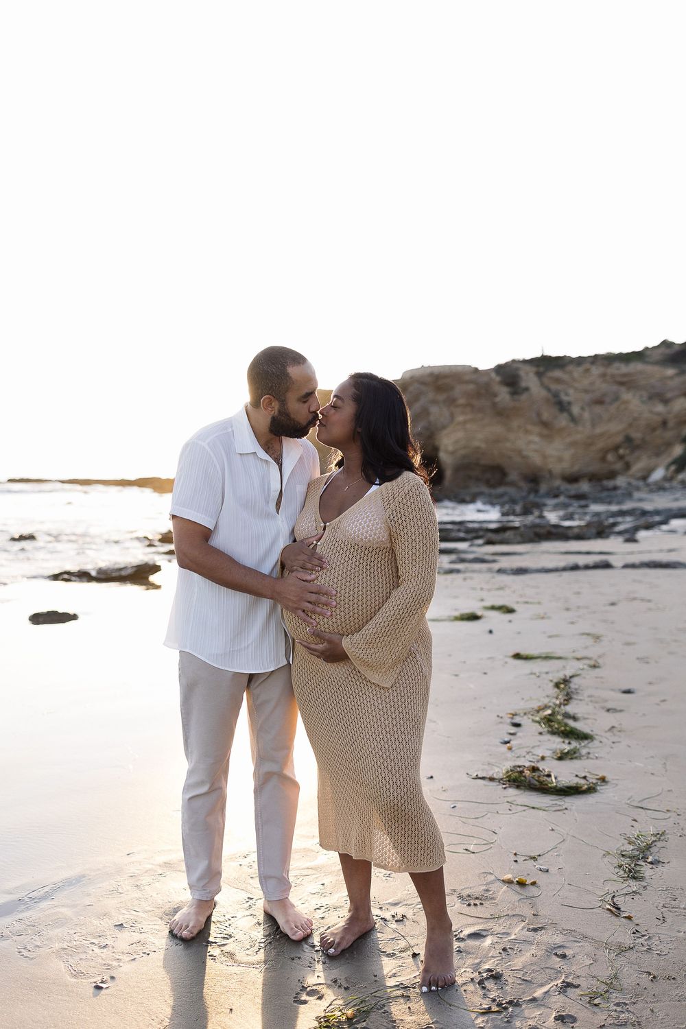 Couple embraces on beach during sunset maternity photoshoot with coastal cliffs in background.