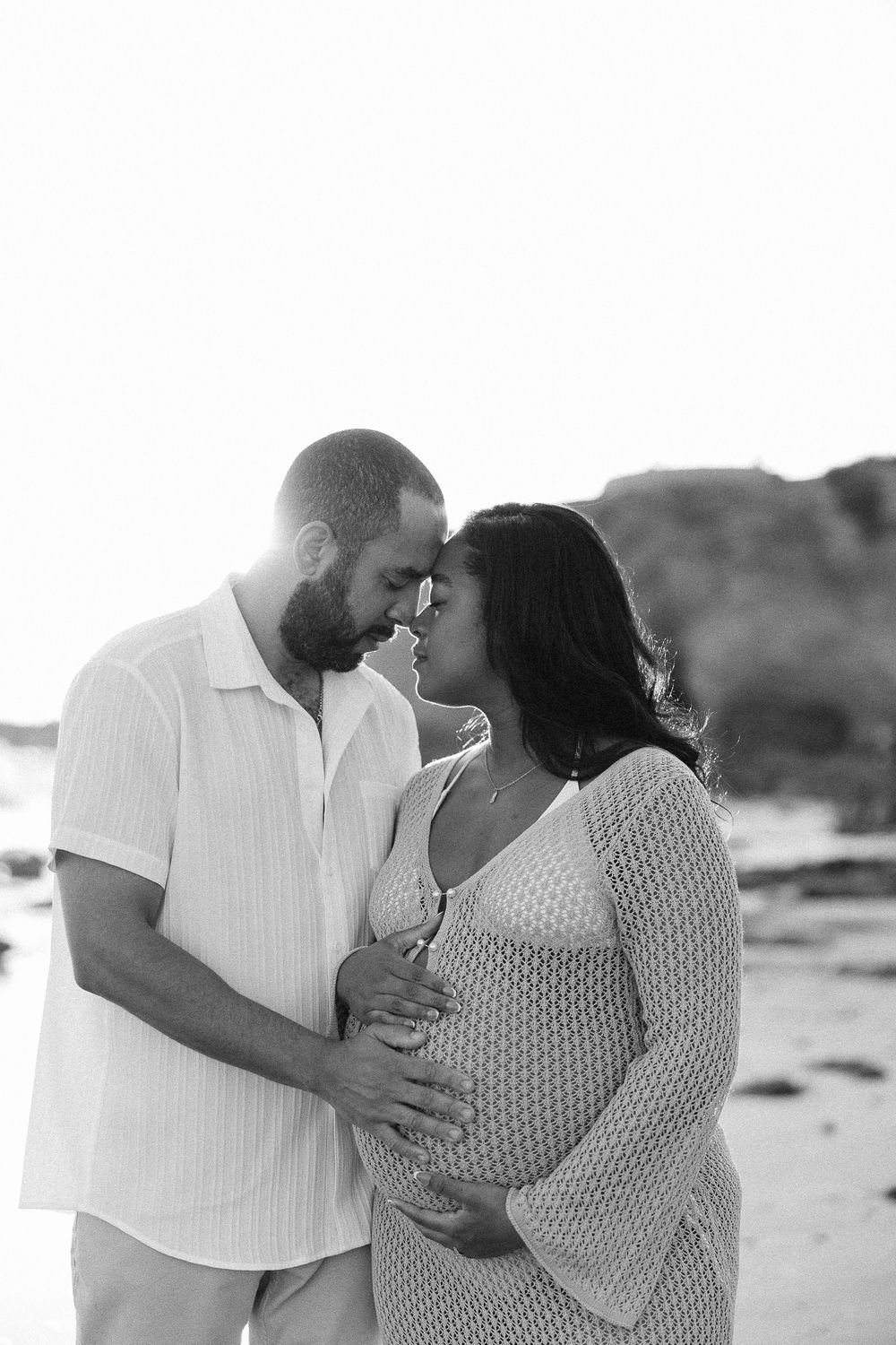 Black and white beach maternity photo of a couple in white outfits sharing an intimate moment at sunset.