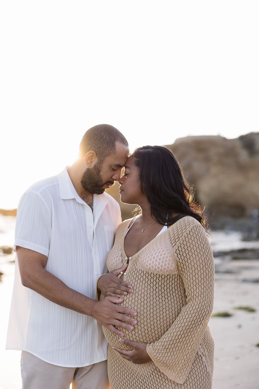 Expecting couple shares intimate moment during sunset maternity photoshoot on rocky beach.