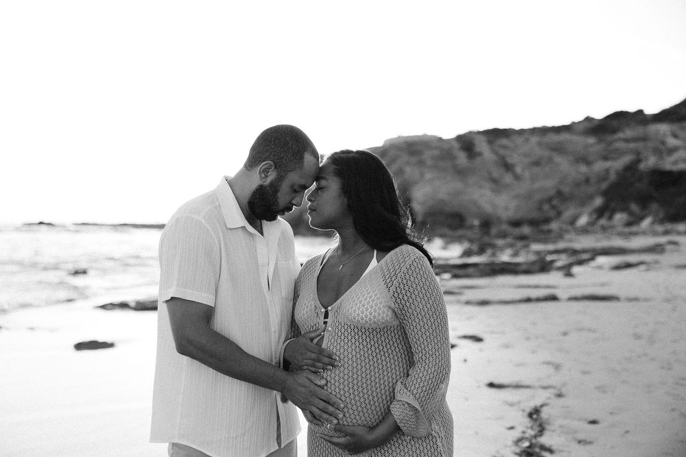 A romantic couple embraces on a scenic beach with rocky cliffs and waves in the background.