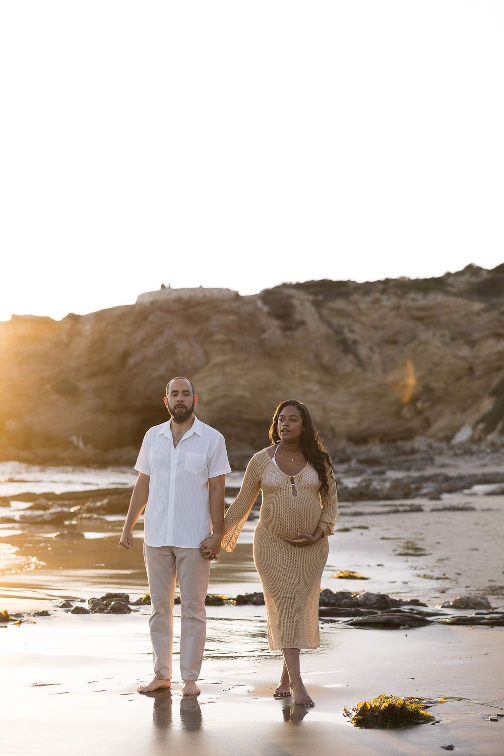 A couple walks hand in hand along a beach at sunset with rocky cliffs in the background.