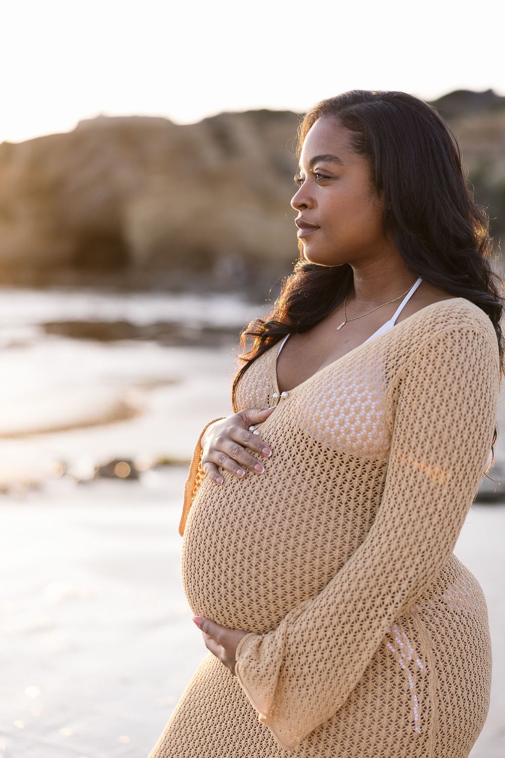 A pregnant woman in a cream knit sweater poses during a beach maternity photoshoot at sunset.