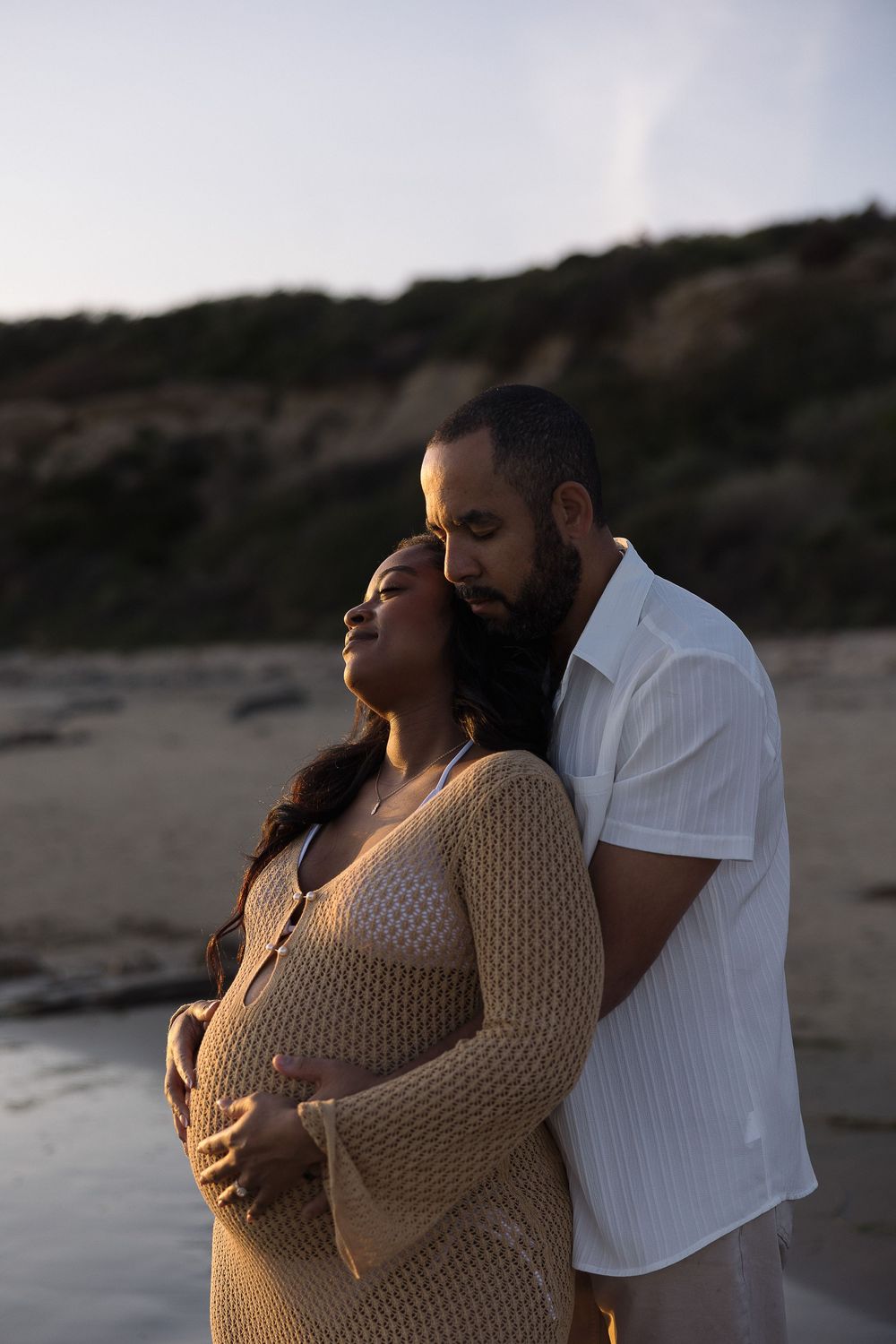 A couple embraces on a beach at sunset as warm light illuminates them against a dark shoreline backdrop.