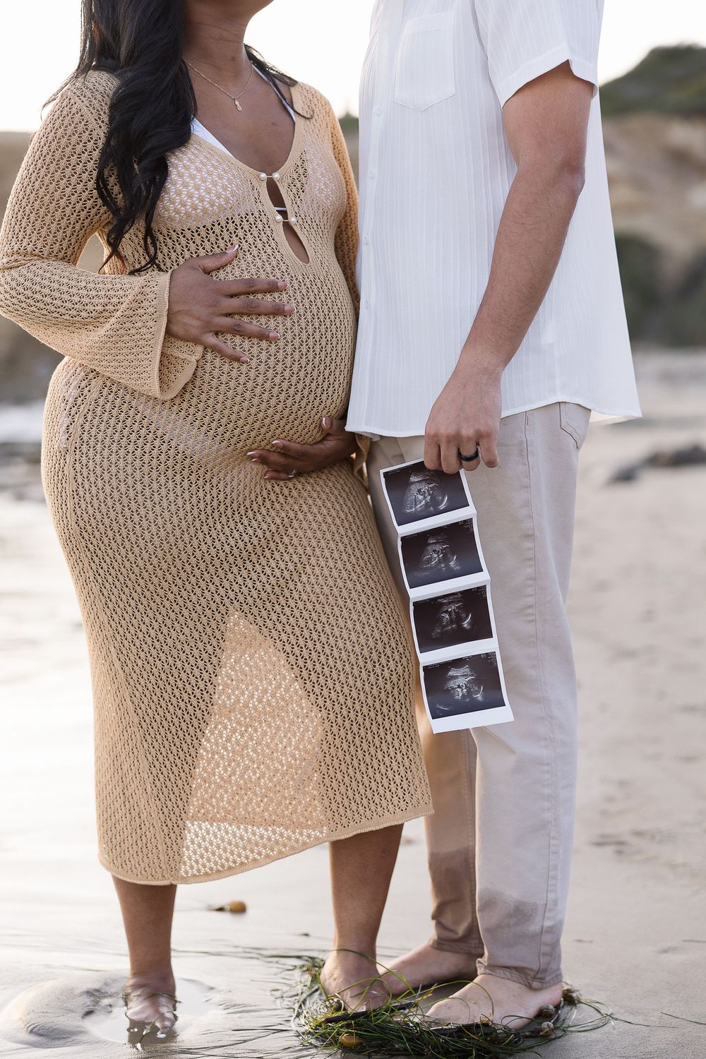 Expecting couple on beach holding ultrasound photos while wearing beige knit dress and white casual outfit.