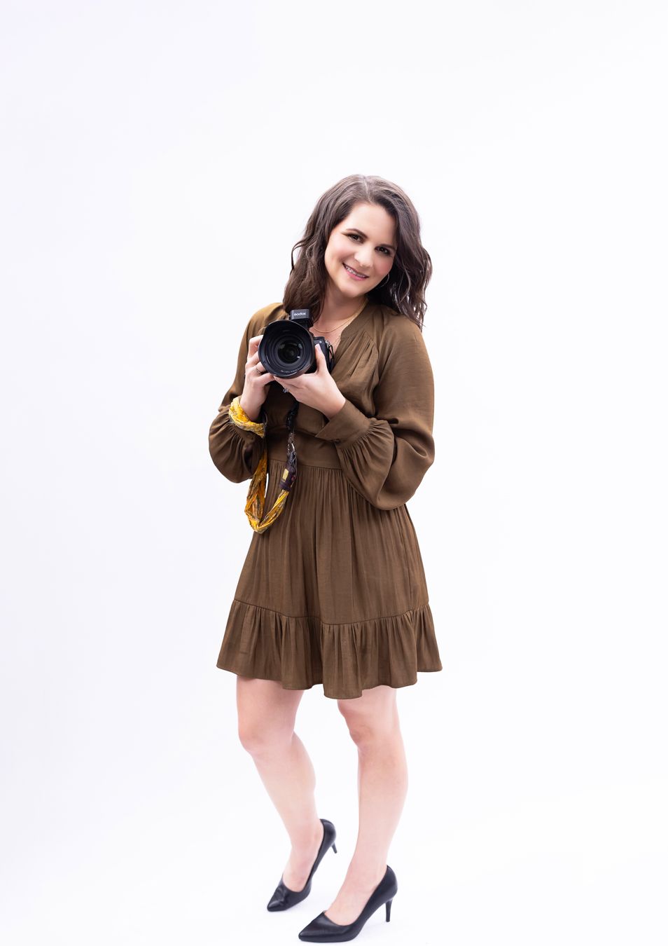 A person in a brown dress holding a camera wearing black heels stands against a white background.