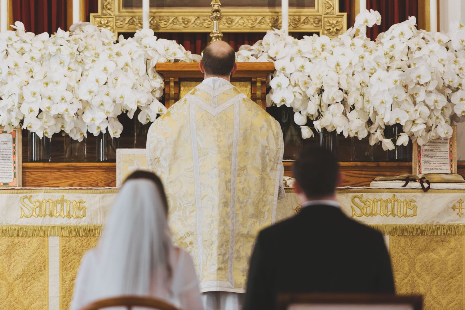 Wedding ceremony view from behind showing bride and groom facing priest at altar decorated with white orchids during Latin Mass.