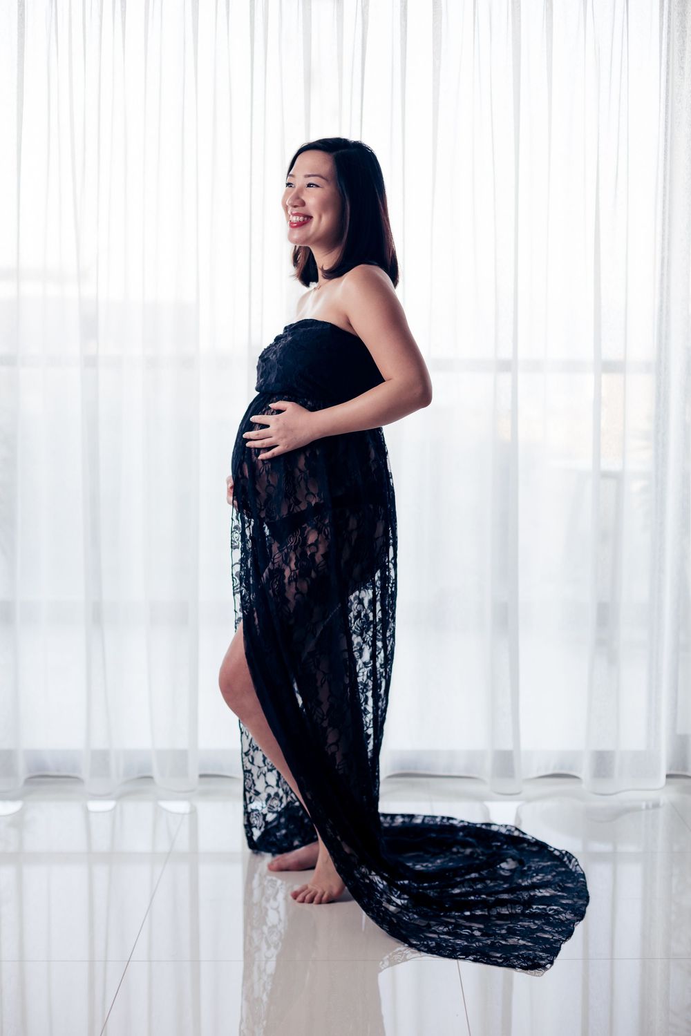 Figure wearing a black lace outfit poses elegantly against white curtains in a maternity photoshoot.