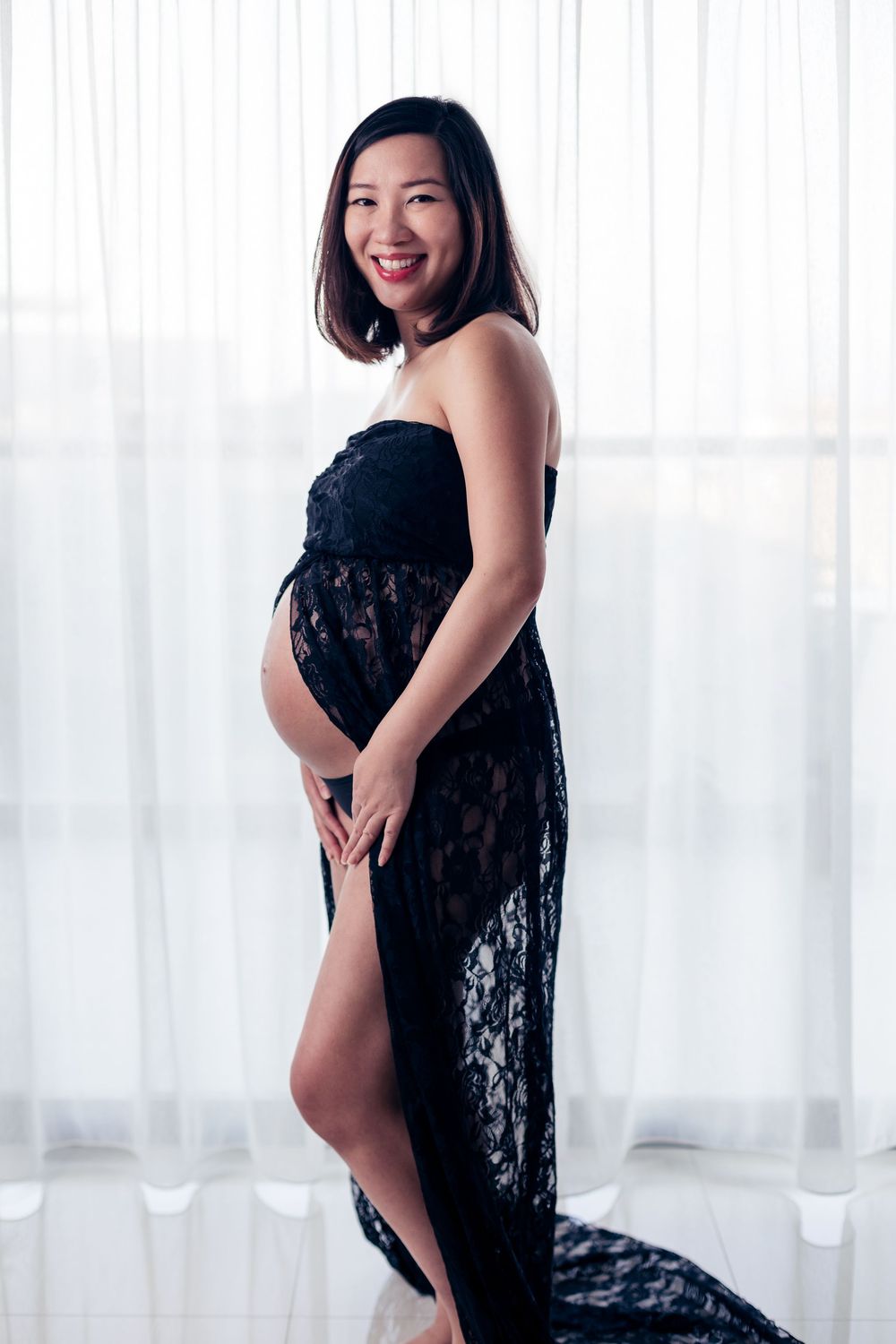 An expectant mother poses in an elegant black lace dress near sheer white curtains during a maternity photoshoot.