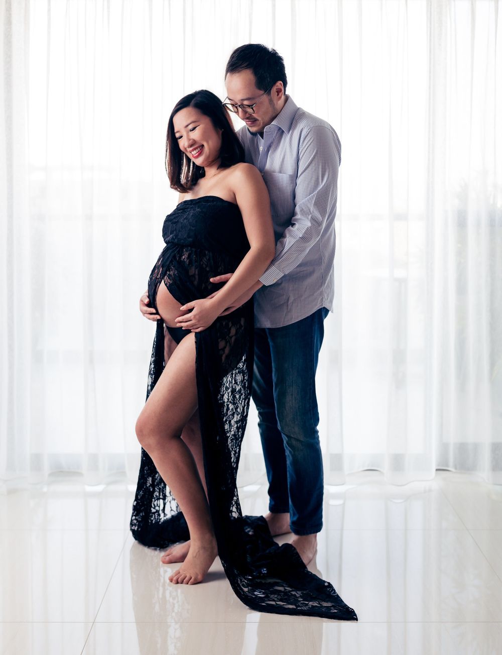 A couple shares an intimate moment in a bright room as they pose for a maternity photo wearing black lace and casual attire.