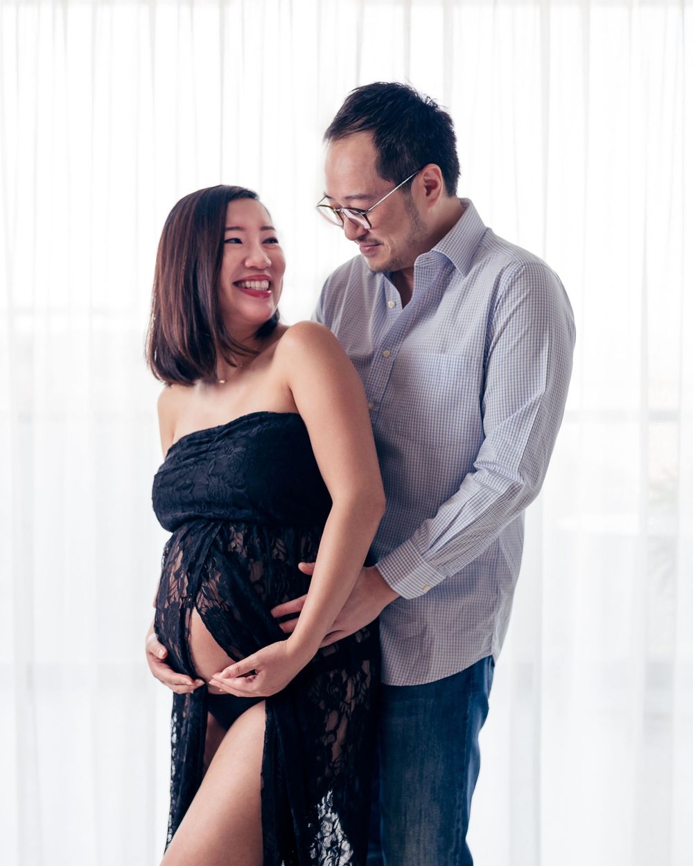 A couple in formal attire stands close together in a bright studio setting with romantic lighting.