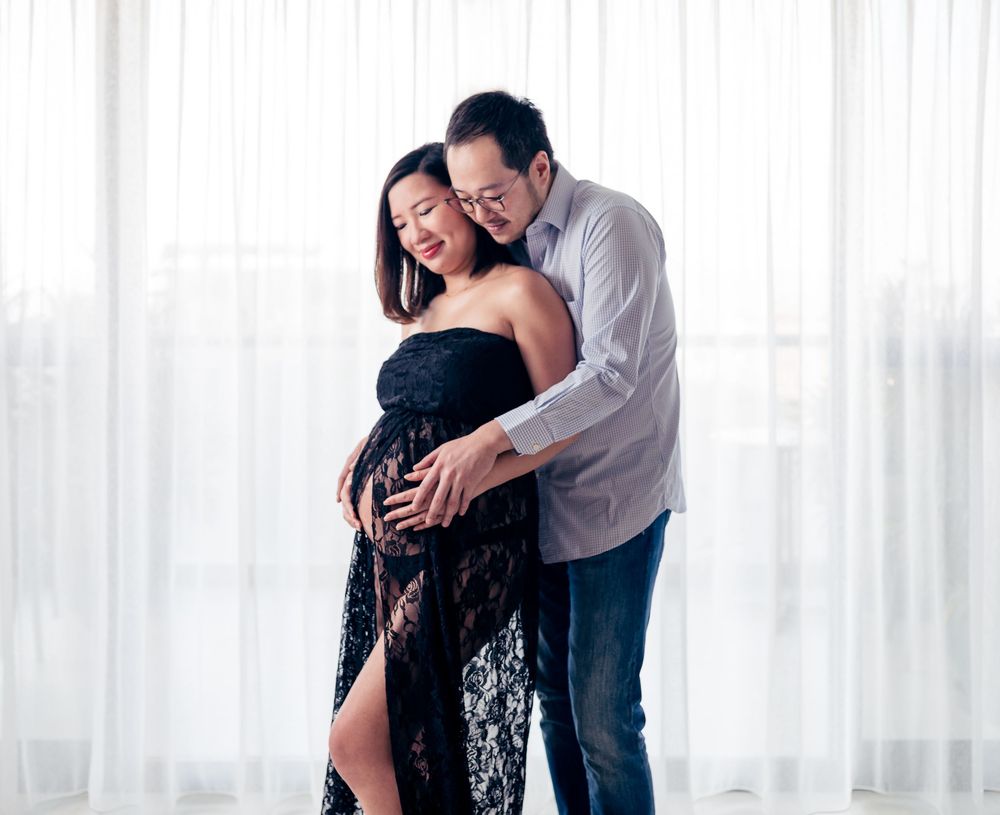 A couple embraces tenderly in front of sheer white curtains in a bright interior space.