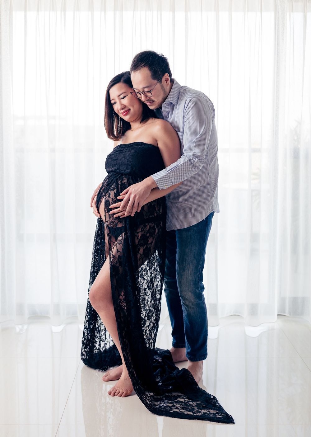 A maternity photo of a couple embracing against white curtains, with the woman wearing a flowing black lace gown.