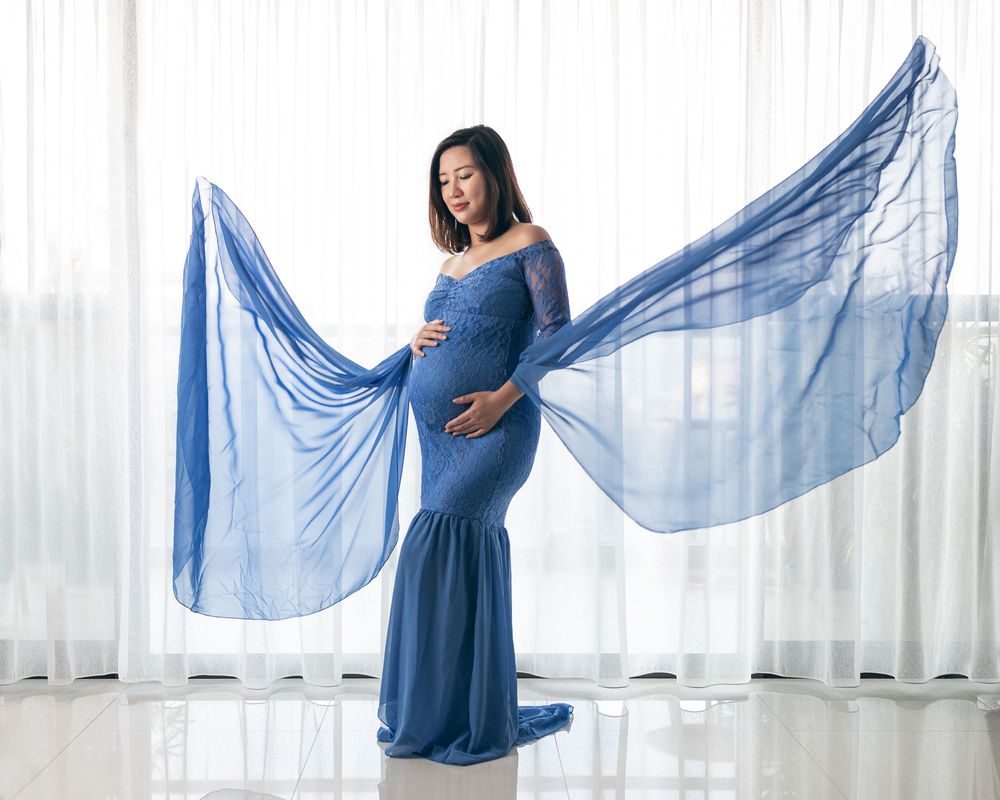 Flowing blue maternity dress with sheer fabric draping elegantly against white curtain background in studio setting.