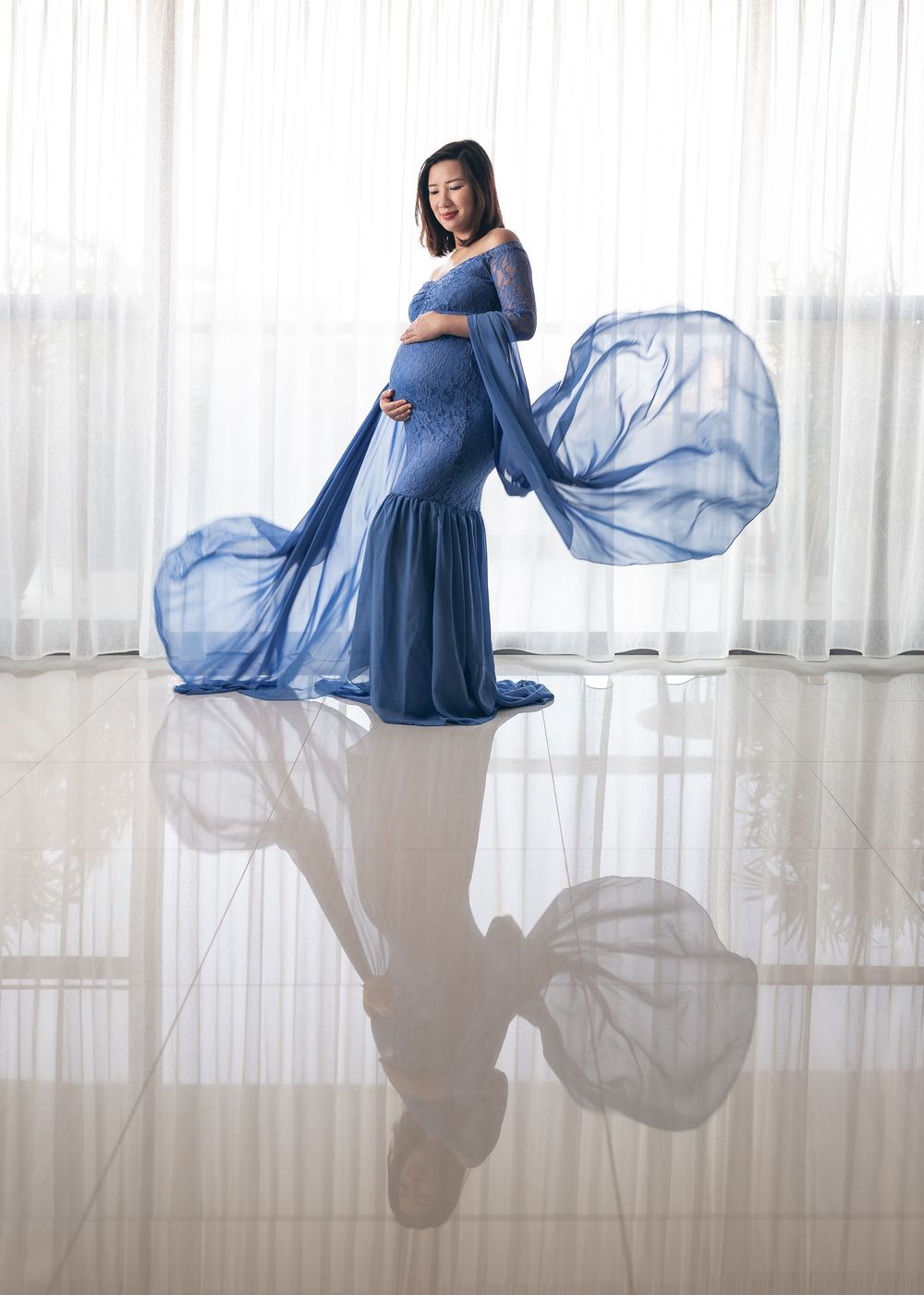 Expectant mother poses in fitted blue gown with flowing sheer fabric creating elegant reflection on glossy floor.