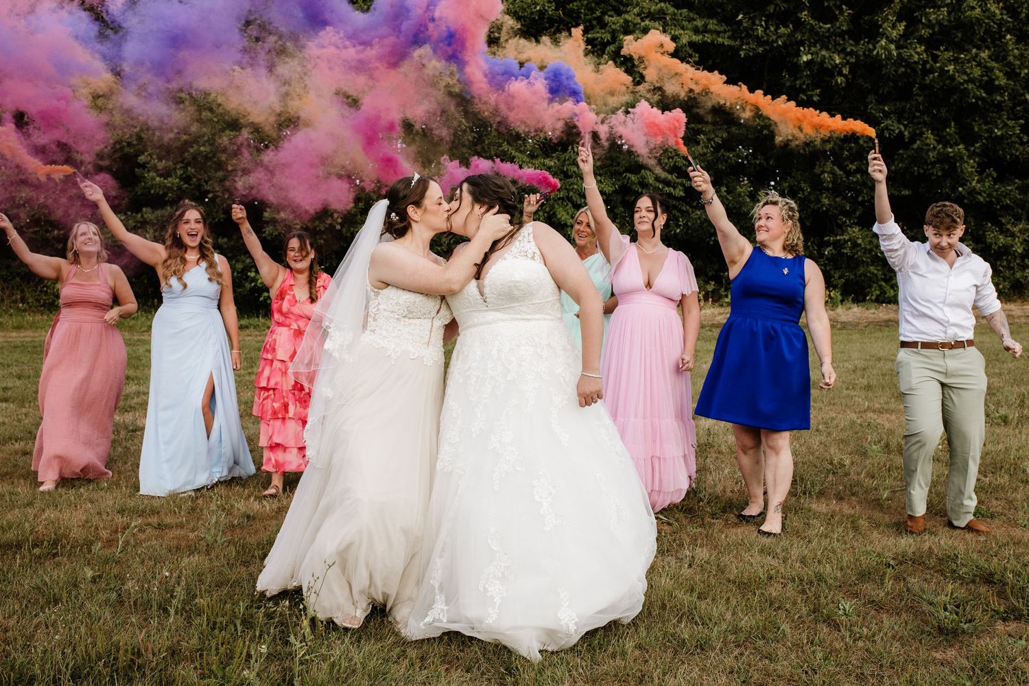 Fun wedding party in matching tan pants and green vests playfully lift someone wearing a white dress in a grassy field.