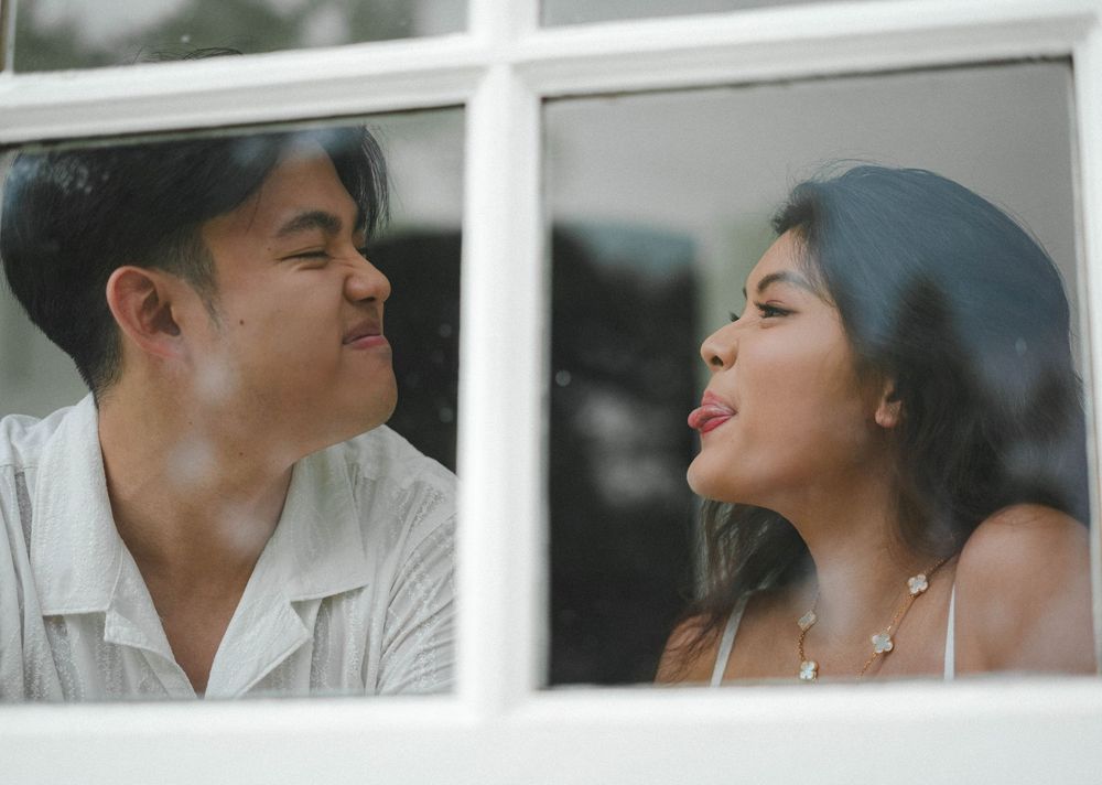 A couple playfully makes faces at each other through a window pane, creating a lighthearted romantic moment.