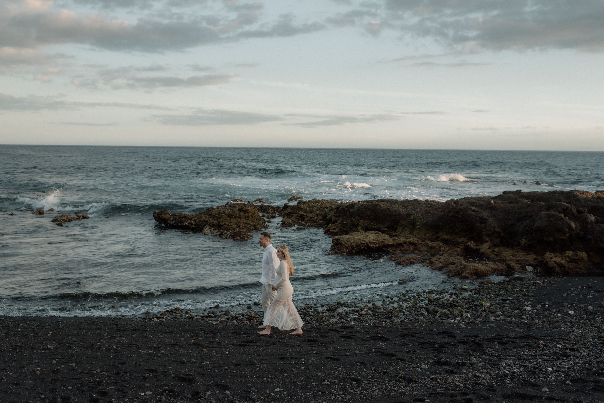 A pregnant woman and in a white dress, stands on a black volcanic beach with rocky coastline and crashing waves in the background.