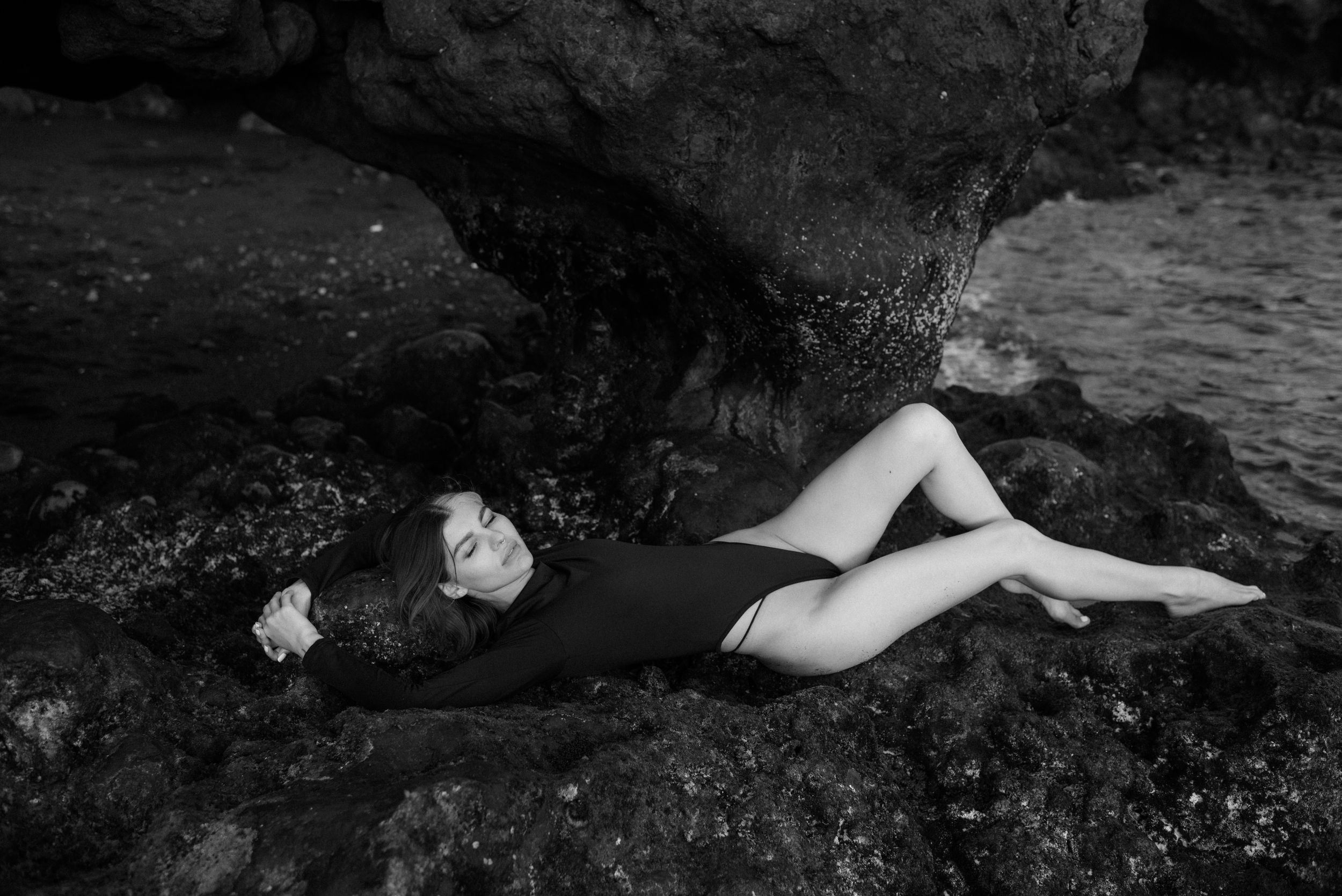 A black and white artistic portrait of a woman against dark rocky formations near water in Tenerife.
