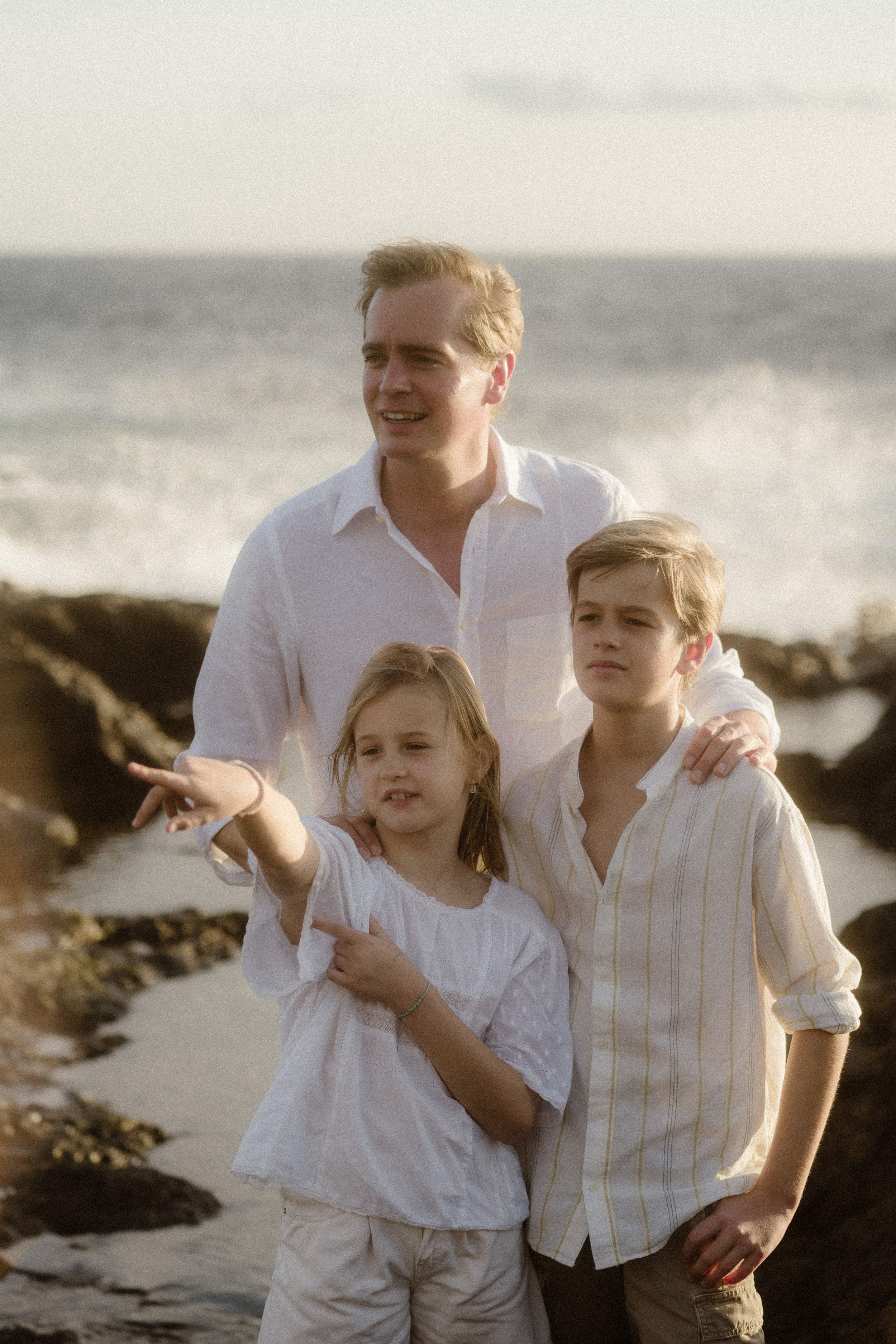 A family wearing white clothing poses together on a rocky beach at sunset with ocean waves in the background.
