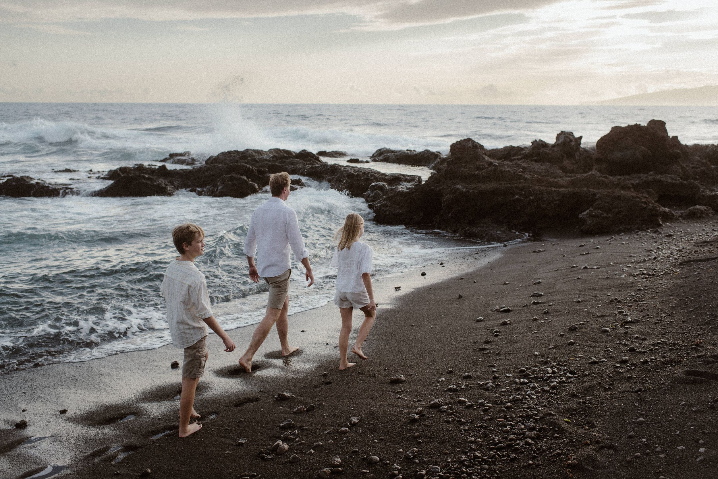 Family in white outfits walking along scenic black sand beach with volcanic rocks and ocean waves at sunset.