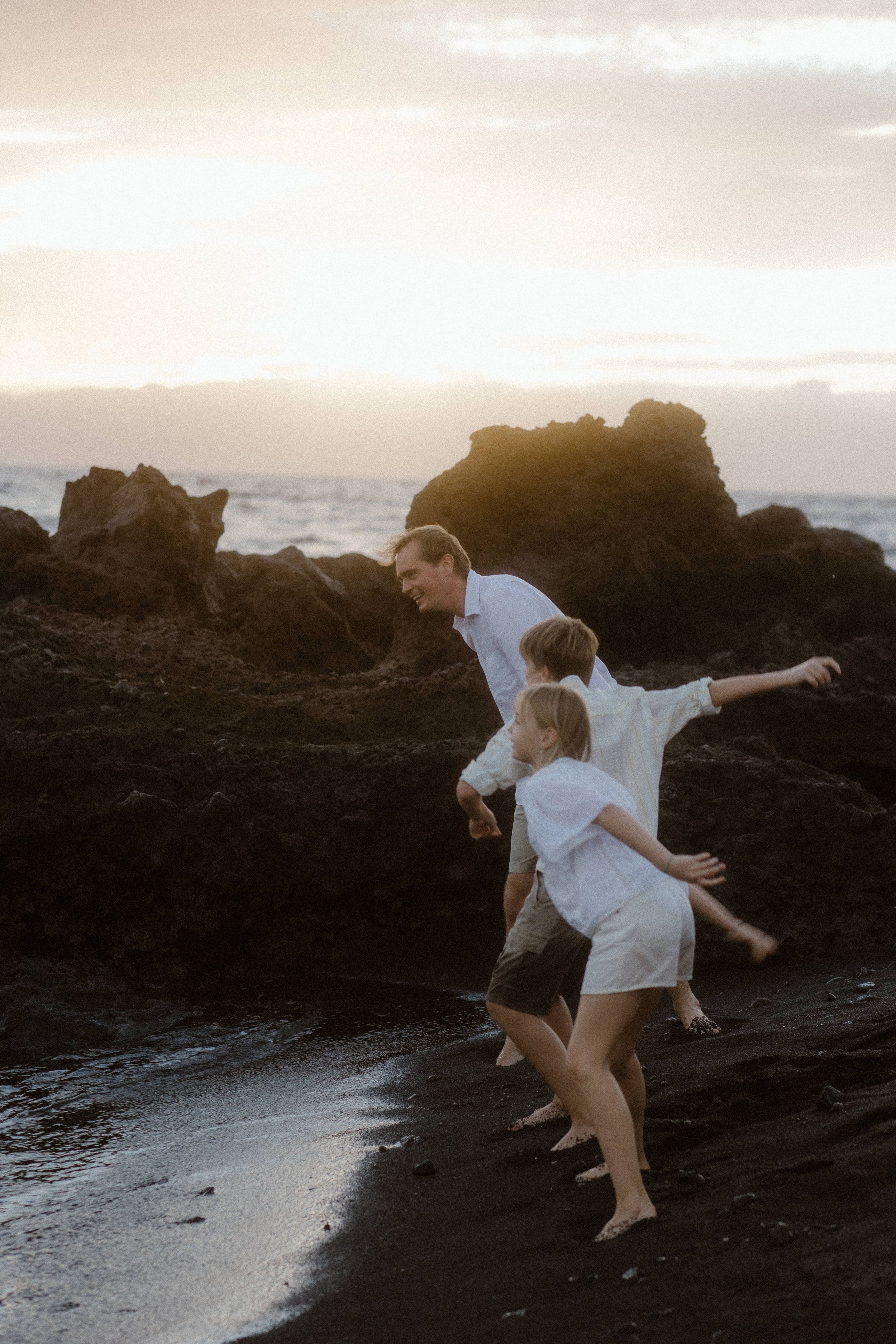 Two people in white outfits leap joyfully on dark rocky beach at sunset with ocean waves and cliffs in background.