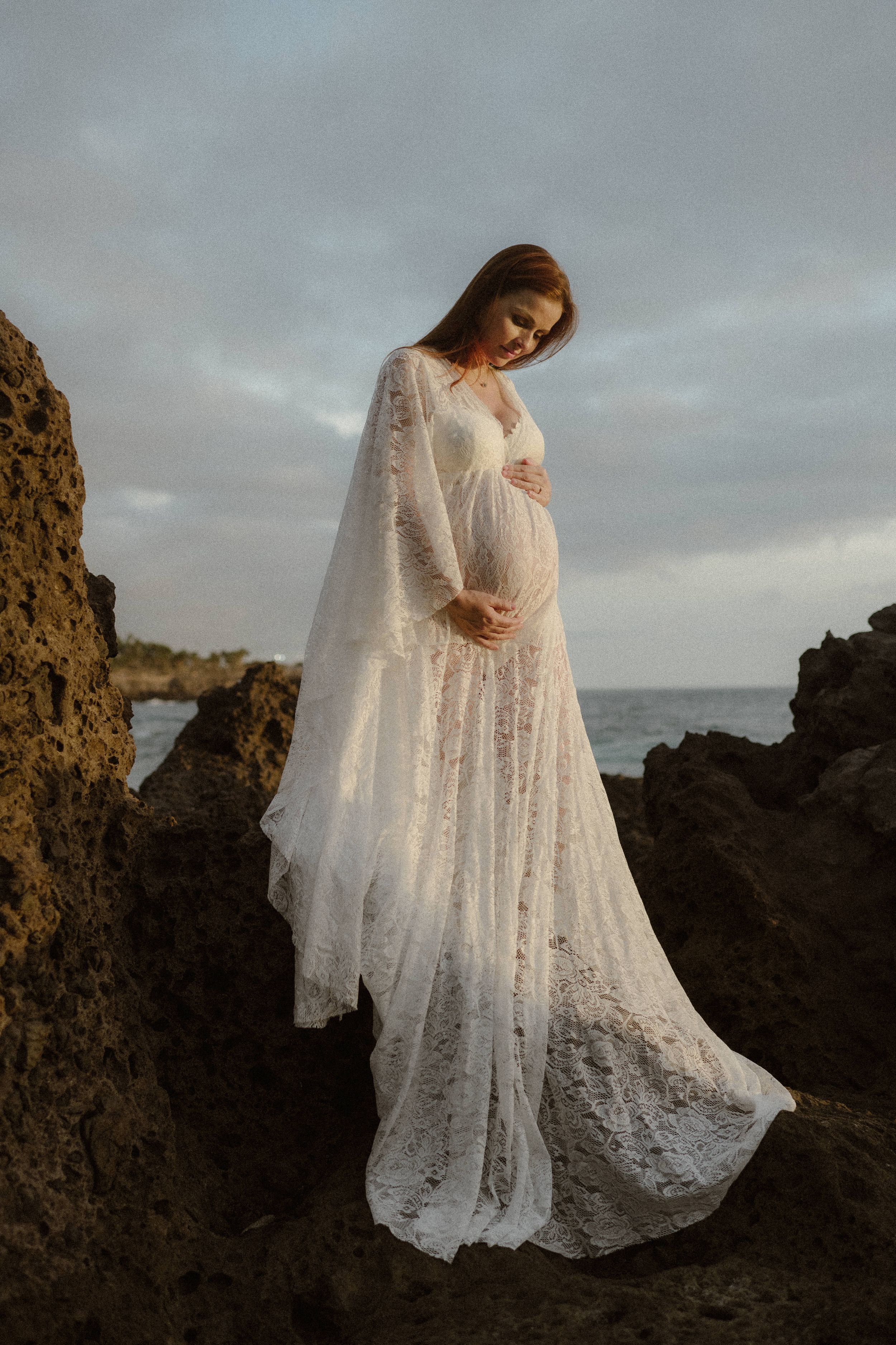 A pregnant woman in a flowing white lace dress and cape stands on rocky coastal cliffs at sunset.